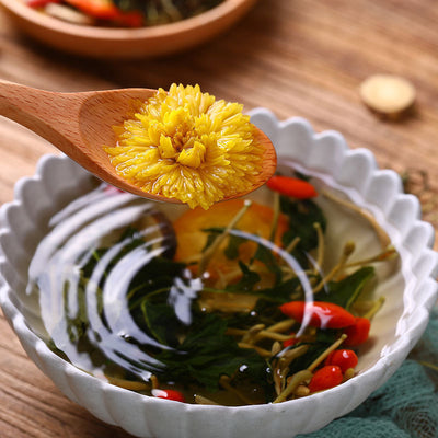 Bright yellow chrysanthemum flower on a wooden spoon above a bowl of honeysuckle chrysanthemum tea with herbs